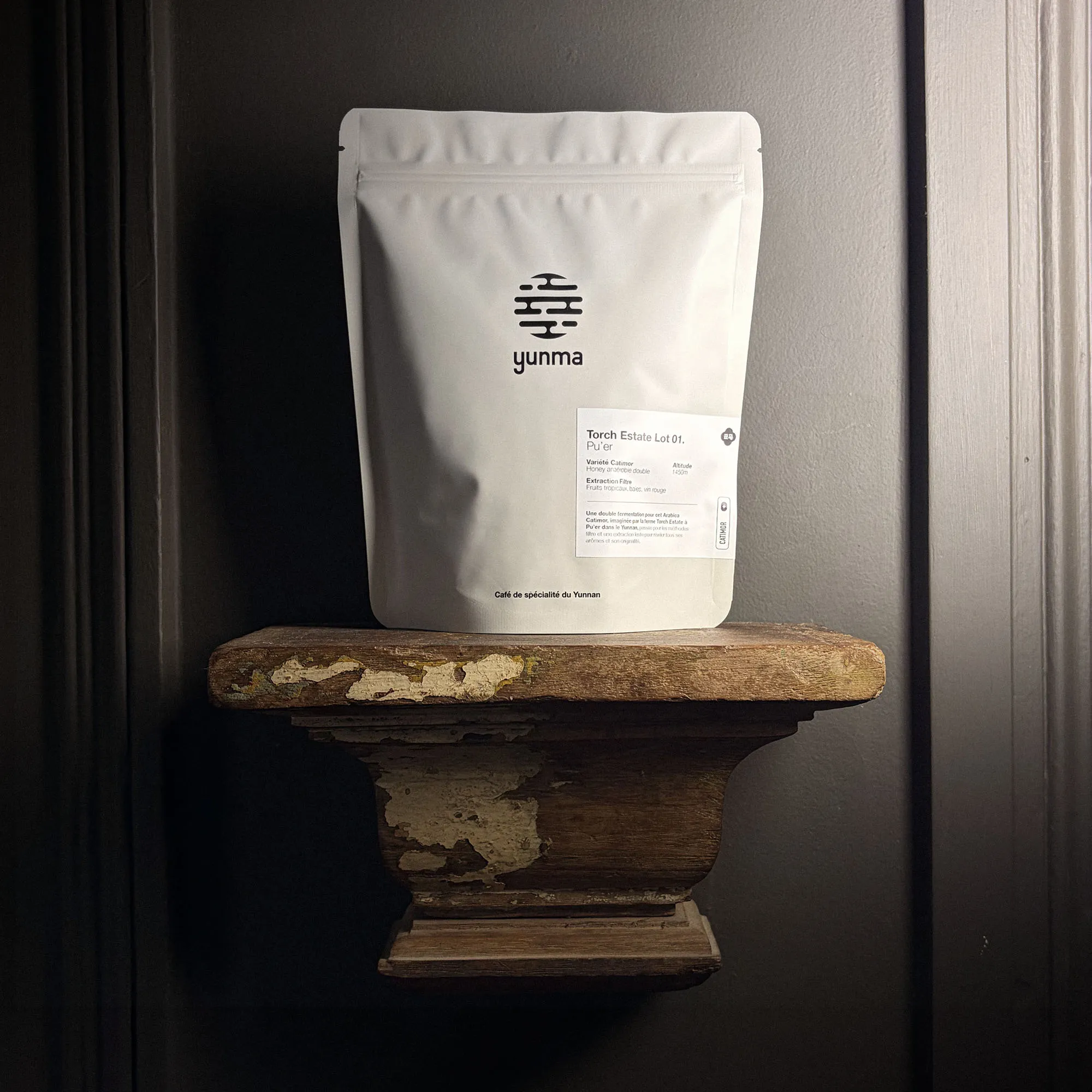Sachet de café Yunma Torch Estate Lot 01, 200 g de grains du Yunnan, honey anaérobie double, posé sur une console de bois patinée devant une porte anthracite